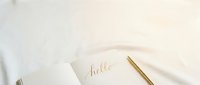 Open notebook with gold hello calligraphy script and elegant golden pen on soft white fabric, minimal flat lay for lifestyle blogging or stationery branding.