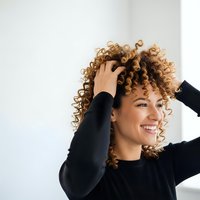 Happy young mixed-race woman with curly hair touching head and smiling in black sweater against white background for lifestyle and wellness concepts.