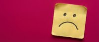 Yellow sticky note with sad face drawn on it against bright pink background expressing disappointment and negative emotions for workplace communication.