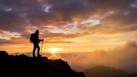 Silhouette of hiker with trekking poles standing on mountain peak at golden sunset, gazing at dramatic sky and distant valleys below.