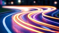 Long exposure photograph of glowing light trails on winding road at night creating vibrant streaks of orange, purple and blue colors against dark background.