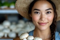 Young Asian woman in straw hat smiling while holding fresh white mushrooms, natural lighting highlights her warm expression and fresh farm produce.
