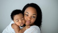 African American mother in white lace dress holding sleeping baby in white outfit against light gray background, tender moment of maternal love and bonding.