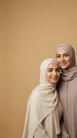 Two young Middle Eastern women wearing hijabs embracing and smiling against beige background for lifestyle and friendship concepts.