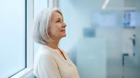 Senior woman with white hair, Caucasian ethnicity, wearing beige blouse, gazing thoughtfully out window in modern office setting. Contemplative professional portrait.