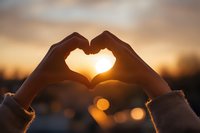 Hands forming heart shape silhouette against golden sunset sky with warm bokeh lights creating romantic atmosphere for love and connection concepts.
