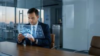 Mature male professional in dark business suit examining holographic digital interface displaying network connections and data visualization in modern office with city skyline view.