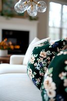Cozy living room interior with white sofa and emerald green floral throw pillows, modern brass chandelier with glass globes, and fireplace in background.