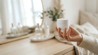 Woman holding white cosmetic jar with cream or skincare product on vanity table with mirror and plants for beauty, wellness, and personal care advertising.