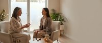 Two women having friendly conversation in modern living room with natural light and plants creating warm atmosphere for lifestyle content.