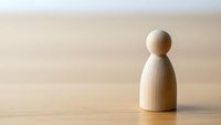 Single wooden peg person figure standing alone on natural wood surface with soft lighting for minimalist design concepts and business presentations.