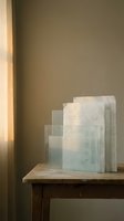 Stacked translucent glass and frosted acrylic sheets arranged on wooden table with warm sunlight casting soft shadows through window, suitable for design and architectural concepts.