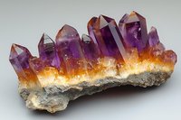 Raw amethyst crystal cluster with vibrant purple points emerging from orange and white crystalline base, displaying natural geometric formations and color gradients.