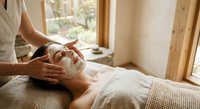 Young woman receiving relaxing facial massage treatment at spa with natural lighting and wooden interior elements for wellness and self-care.