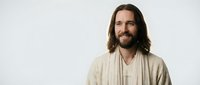 Young Caucasian man with long brown hair and beard wearing beige robes smiling warmly against white background for spiritual and religious content.