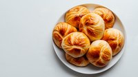 Fresh homemade butter rolls on white ceramic plate, golden brown baked pastries with flaky layered texture and glossy surface, shot from above on light background.
