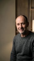 Confident senior Caucasian man with gray stubble smiling warmly indoors, wearing a gray sweater in soft natural light.