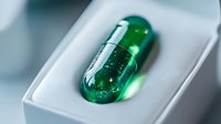 Green transparent capsule with glittering particles inside resting in white packaging, representing pharmaceutical innovation or nutritional supplement for health industry.