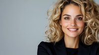Young Caucasian businesswoman with curly blonde hair and bright smile wearing black blazer against gray background, professional corporate headshot style portrait.