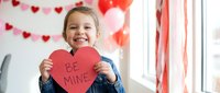Happy young girl holding red heart-shaped card with Be Mine text for Valentine's Day celebration at home with festive decorations and balloons.