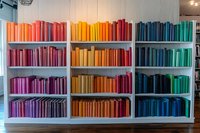 Books arranged by color on white wooden shelves creating rainbow spectrum from pink through yellow to blue. Modern library interior with color-coordinated organization.