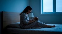 Young Caucasian mother sitting on bed at night, cradling newborn baby in dim blue light, tender moment of late-night feeding and bonding.
