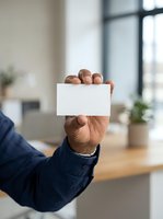 Mature male professional with medium skin tone holding blank white card in modern office environment with wooden desk and natural light from windows.