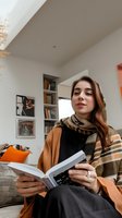 Young Mediterranean woman reading book at home, wearing casual autumn outfit with orange cardigan and scarf. Cozy interior with bookshelf and natural lighting creates relaxing atmosphere.