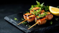 Grilled salmon skewers with fresh herbs and lemon wedges served on black stone plate against dark background, dramatic food photography with natural lighting.