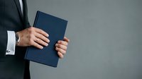 Businessman in dark suit holding navy blue notebook or journal against neutral gray background, cropped torso view with copy space for corporate messaging.
