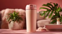 Rose gold stainless steel travel mug with flip lid on pink surface surrounded by tropical plants creating modern lifestyle aesthetic for beverages and hydration.