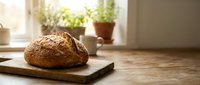 Freshly baked artisan bread loaf on wooden cutting board in sunlit kitchen with potted herbs on windowsill, perfect for bakery and homemade cooking concepts.