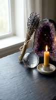 Spiritual wellness setup with amethyst geode, dried lavender bundle, crystal ball and burning candle on wooden surface near window for meditation and healing.