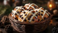 Fresh hot cross buns with raisins and white icing glaze in rustic wicker basket, decorated with pine cones and warm candlelight for Easter celebration.