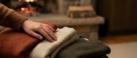 Young child with pale skin resting hand on stack of folded knit sweaters in warm earth tones, cozy home interior setting with fireplace glow. Perfect for lifestyle, family, comfort, and seasonal content.