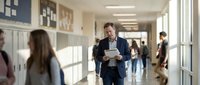 Middle-aged Caucasian businessman in navy suit walking through bright school hallway with documents, educational administration concept for corporate training.