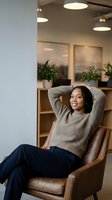 Relaxed African American woman with braids sitting in leather armchair at modern office, hands behind head, smiling confidently in cozy workspace.