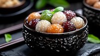 Colorful sweet rice balls in black ceramic bowl garnished with mint leaf, traditional Asian dessert served on dark stone background with decorative elements.