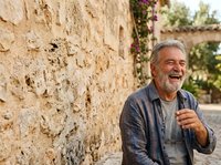 Mature man with gray hair and beard, Mediterranean ethnicity, wearing casual blue shirt, standing against weathered stone wall with ivy, outdoor portrait for lifestyle and travel content.