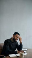 Man of African descent experiencing work-related stress, holding head while seated at desk with notebook and coffee mug, suitable for business, mental health, and workplace wellness content.