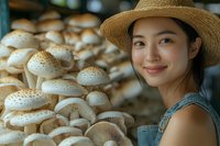 Young Asian woman in straw hat and denim overalls smiling next to fresh organic mushrooms growing in rows at indoor farm. Natural lighting highlights sustainable agriculture.