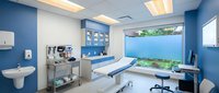 Modern medical examination room with blue walls, white equipment, dental chair, and large window overlooking garden for healthcare facility design.