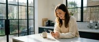 Woman with long brown hair, wearing a beige ribbed sweater, sitting at a white desk in a modern kitchen with black-framed windows, holding a smartphone and smiling while working remotely.