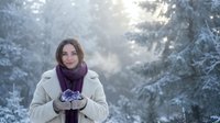 Young woman holding snow in hands wearing winter coat and purple scarf in snowy forest with sunlight rays filtering through trees during cold season.