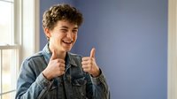 Happy teenage boy with curly hair giving thumbs up gesture in denim jacket, smiling with positive attitude in modern indoor setting for youth lifestyle content.