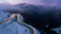 Aerial view of winding mountain road with vehicle light trails at dusk, snow-covered landscape with forested valleys and peaks, winter travel and transportation concept.