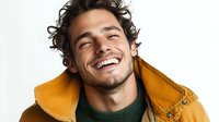 Young Mediterranean man with curly hair wearing yellow jacket and green sweater laughing with genuine joy against white background, expressing happiness and positive emotions.
