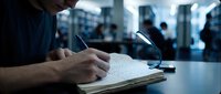 Man in business casual attire writing notes in notebook at desk during corporate meeting or office presentation with blurred colleagues in background.