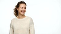Young woman with curly brown hair and fair complexion wearing a cream-colored ribbed sweater, smiling against white background for portrait and lifestyle use.