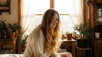 Young Caucasian woman with long wavy blonde hair in cozy beige sweater sitting by sunny window in rustic home interior with plants and warm natural lighting.